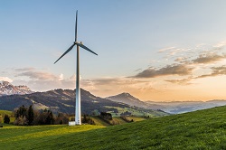 Image of a wind turbine standing in a green field with mountain scenery and a rising sun in the background
